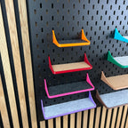 Vier Ablagen mit Rand in Orange, Rot, Pink und Lila untereinander an schwarzer Lochwand