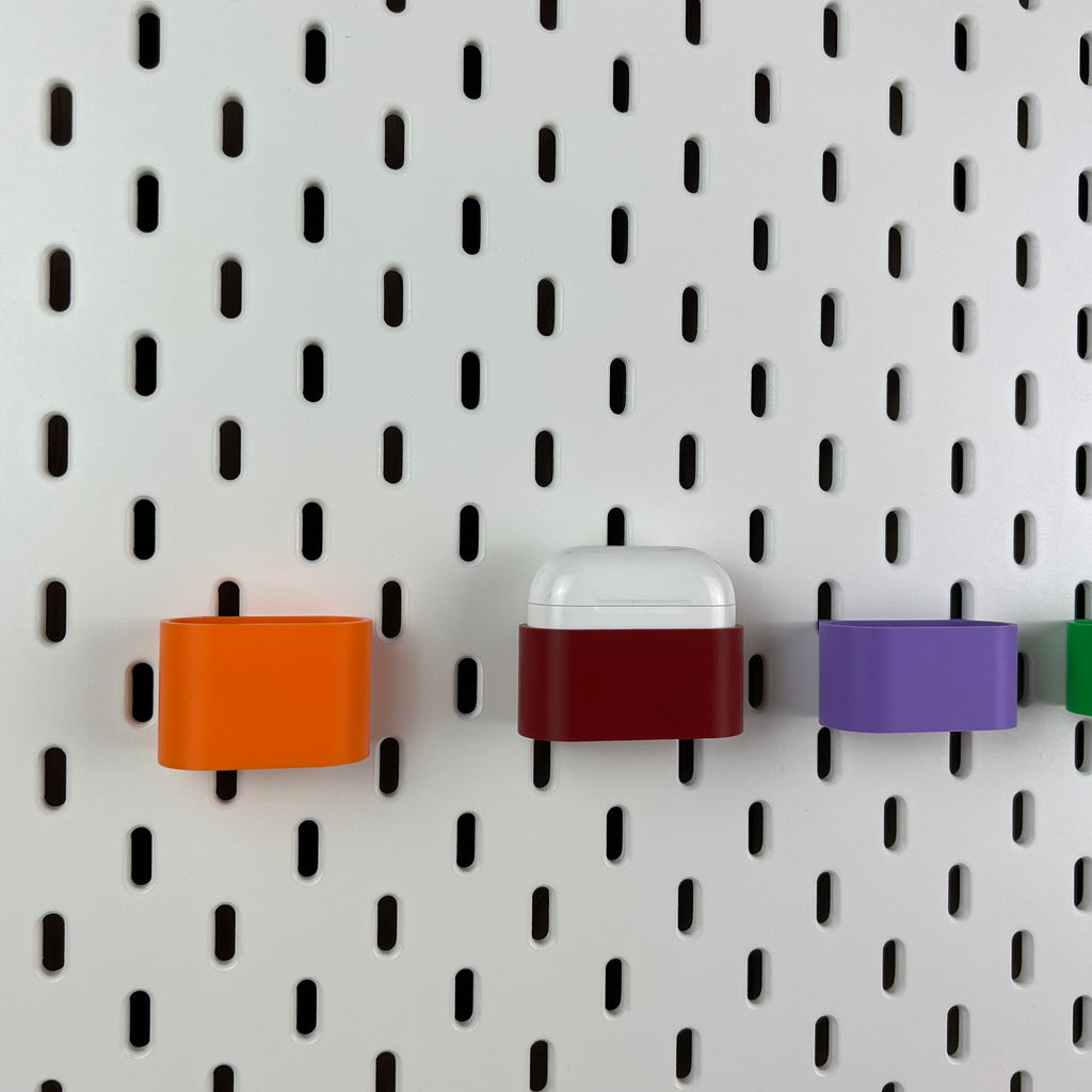 AirPods-Halterungen in Orange, Dunkelrot und Lila an weißer SKÅDIS Lochwand, eine mit AirPods