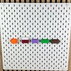AirPods-Halterungen in Orange, Rot, Lila, Grün und Braun an weißer SKÅDIS Lochwand
