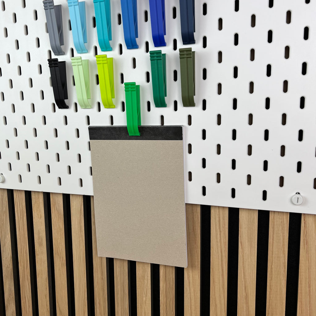 Grüne und blaue SKÅDIS Klipps halten einen Notizblock an der Lochwand vor einer Akustik-Holzwand