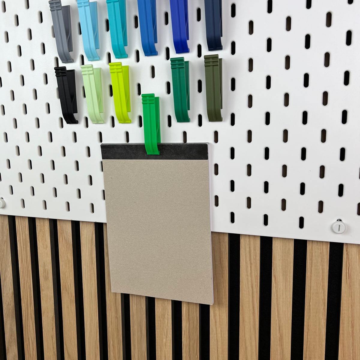 Grüne und blaue SKÅDIS Klipps halten einen Notizblock an der Lochwand vor einer Akustik-Holzwand