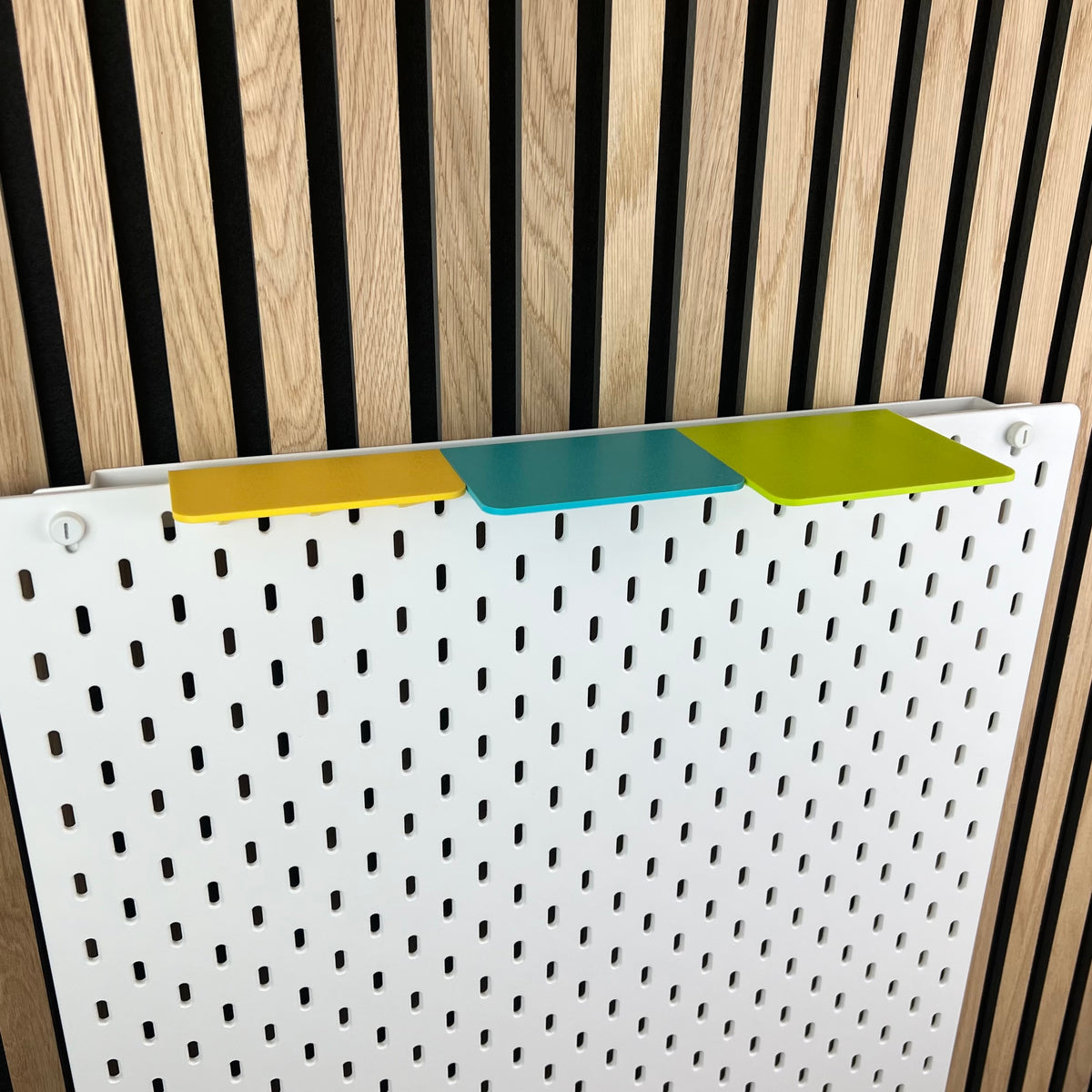 Weiße IKEA SKÅDIS Lochwand mit drei bunten TOP-Ablagen vor Akustikpaneel aus Holz